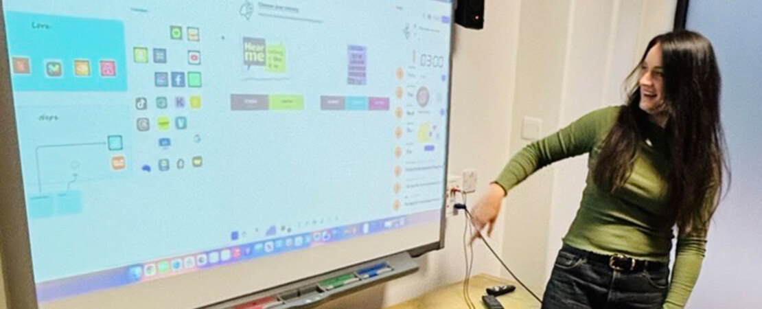 Smiling woman points and looks at large screen behind her, her body faces the room. The screen displays app icons and colour palettes.