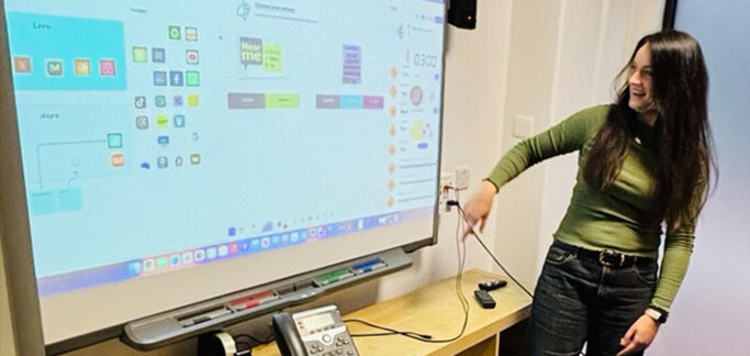 Smiling woman points and looks at large screen behind her, her body faces the room. The screen displays app icons and colour palettes.