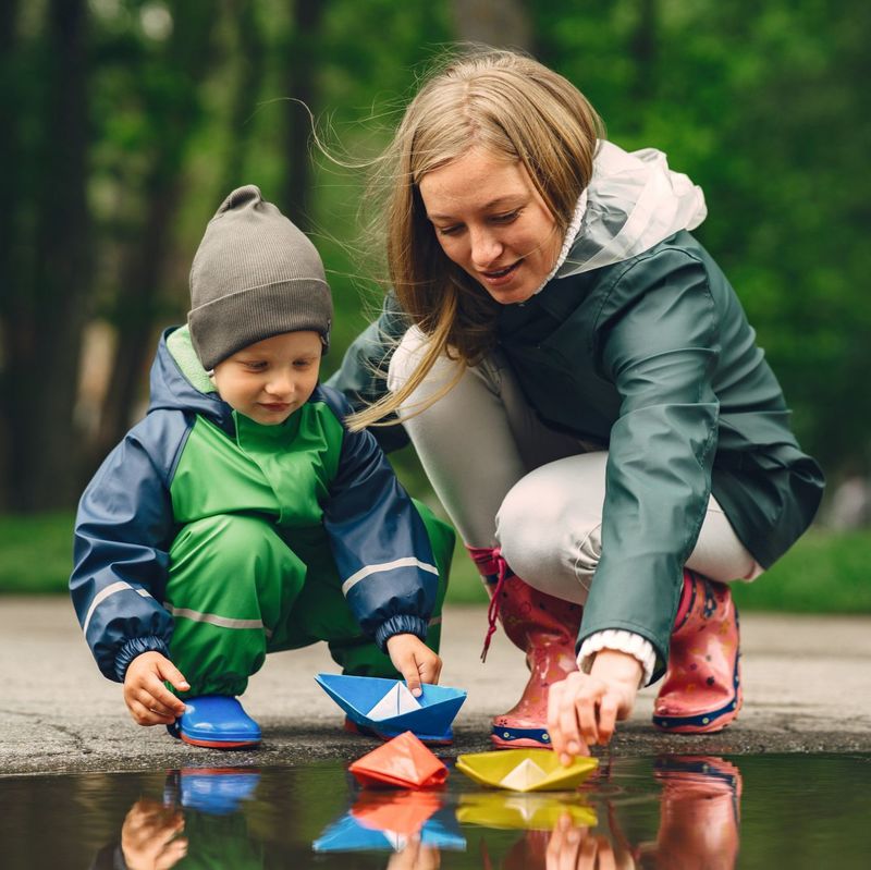 A parent and child playing with paper boats in a puddle