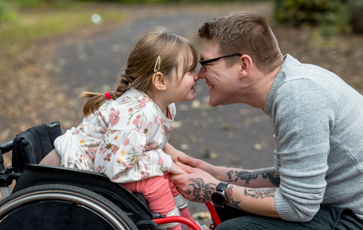 A close up side view of a father and his young daughter who is a wheelchair user